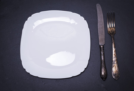 empty white square ceramic plate and iron fork with knife on black background, top view