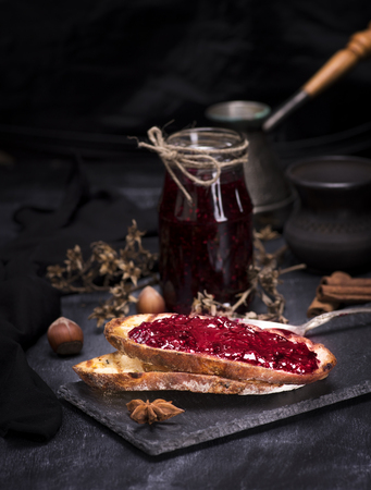 toasts of white bread with raspberry jam on a black surface, behind a glass jar with jam
