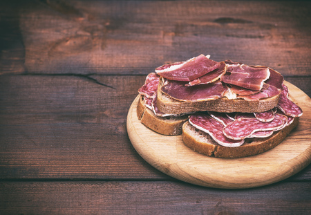 sandwiches with smoked sausage salami and pieces of hamon on a round wooden board, empty space on the leftの写真素材