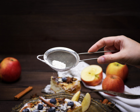 A female hand holds an iron sieve with powdered sugar and sprinkles an apple pieの写真素材