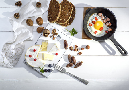 round camembert cheese, pieces of smoked sausage and a round black cast-iron frying pan with a fried chicken egg on a white wooden table, top viewの写真素材