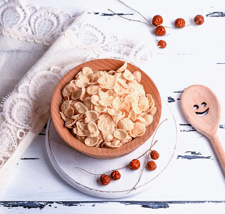 cornflakes in a wooden bowl on a white board, top view, next to a wooden spoon