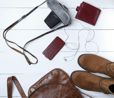 leather brown boots, smartphone in a case with headphones, purse and old vintage camera in a black case with a strap, top view, white wooden backgroundの写真素材