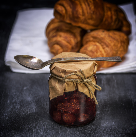 a glass jar with strawberry jam and an iron spoon on a black wooden tableの写真素材