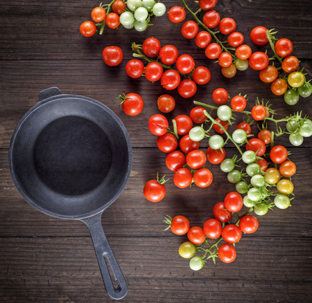 empty black cast-iron frying pan and red cherry tomatoes on a brown wooden table, top viewの写真素材