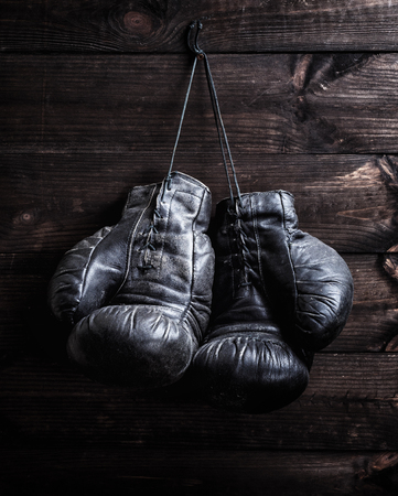 pair of very old shabby black leather boxing gloves hanging on a nail against a brown wooden wallの写真素材