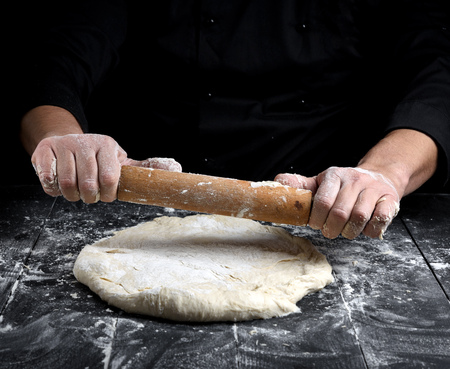 chef in a black tunic rolls a dough for a round pizza on a black table with a wooden rolling pin