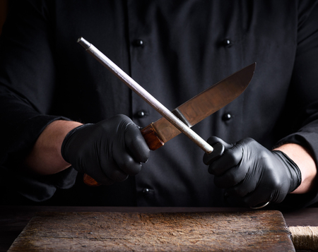 cook in black latex gloves sharpens a knife over a wooden tableの写真素材