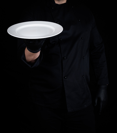 cook in black uniform and black latex gloves holds in his hand a round empty white plate, black backgroundの写真素材