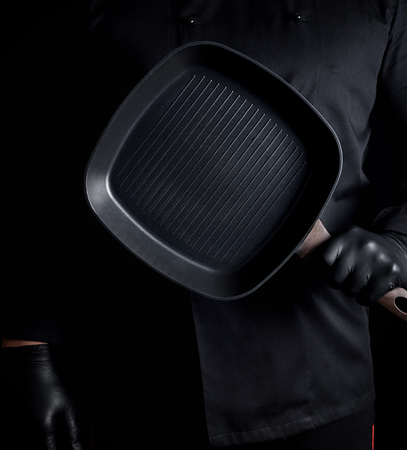 Chef in black uniform and black latex gloves holding an empty square grill pan, black backgroundの写真素材