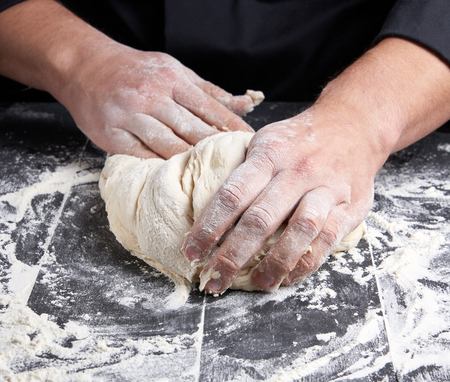 baker kneads white wheat flour dough on a black wooden table, top view