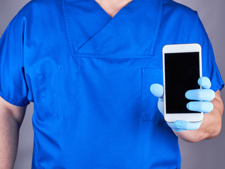 doctor in blue uniform and latex gloves showing a white smartphone with a blank black screen, gray backgroundの写真素材