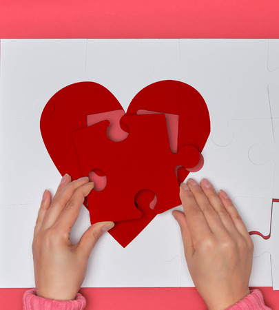 Female hands hold big white puzzles with a piece of red heart, pink background, top viewの写真素材