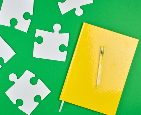 closed yellow notebook and pen, next to it are large empty white paper puzzles, green background, top viewの写真素材