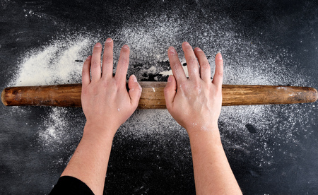 Brown wooden rolling pin in female hands on a black background with white wheat flour, top viewの写真素材