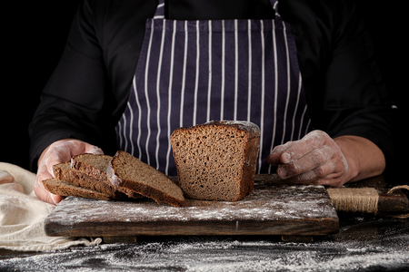 chef in black uniform sliced baked rye bread on a brown wooden board sprinkled with white flour,...