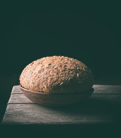 baked round rye-bread bread with sunflower seeds lies on a brown clay plate, black background