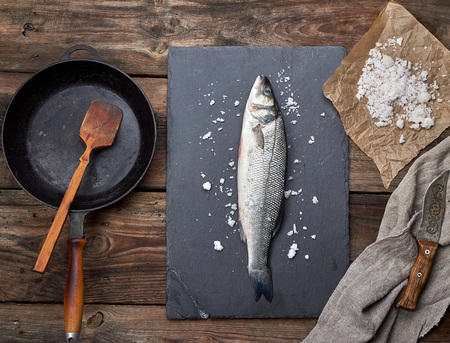 fresh whole sea bass fish on a black graphite board, next to it is an empty round black frying pan on a wooden table, top viewの写真素材