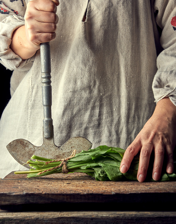woman in a gray linen dress is cutting green leaves of fresh sorrel on a wooden cutting board with an old metal knife, black backgroundの写真素材