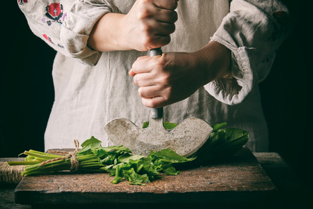 Woman in a gray linen dress is cutting green leaves of fresh sorrel on a wooden cutting board with an old metal knife, black backgroundの写真素材