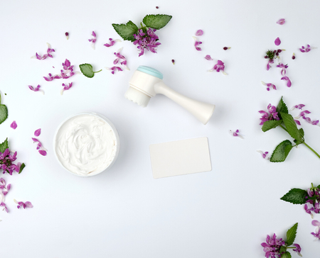 white moisturizing thick cream in a plastic jar, massager and empty paper white business card, white background with flowers and green leaves, top viewの写真素材