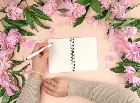 female hand holds in her left hand a white pen over a spiral notebook with empty white pages, next...