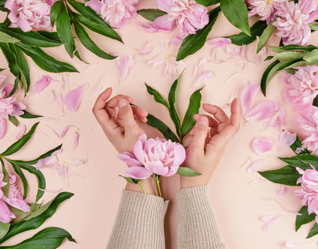 two hands of a young girl with smooth skin and a bouquet of pink peonies on a peach background, top view,  fashionable concept for hand skin care, anti-aging care, spa treatmentsの写真素材