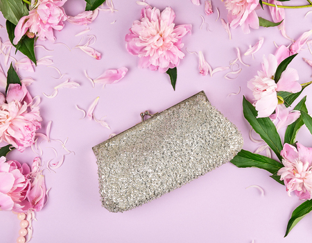 Closed silver women's handbag with sequins for cosmetics on a lilac background with blooming buds of pink peony, top viewの写真素材