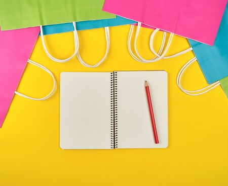 open notebook with blank white sheets and multi-colored paper shopping bags with white handles on a yellow background, flat layの写真素材