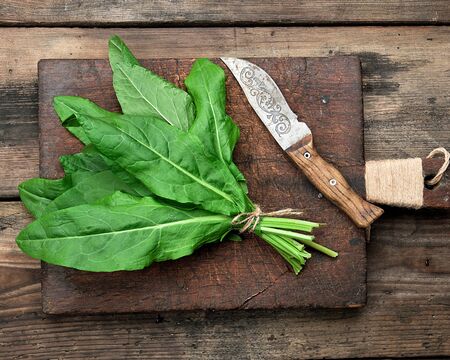 bunch of fresh green sorrel leaves and old brown cutting board on a wooden table, top view