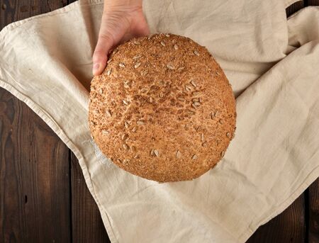 baked round rye bread with sunflower seeds on a beige textile napkin, wooden background, top view