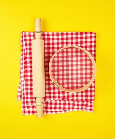 new wooden rolling pin on a red textile napkin and a round sieve for flour, yellow   background, top viewの写真素材