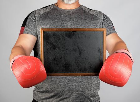 adult athlete in gray uniform and red leather boxing gloves holding a blank black frame, place for an inscription, gray backgroundの写真素材