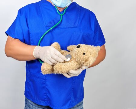 doctor in blue uniform and white latex gloves holding a brown teddy bear, pediatrician listens with...