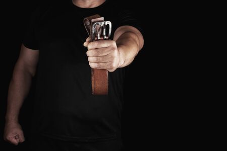 man in black clothes is holding a brown leather belt with a buckle, concept of aggression and domestic violenceの写真素材