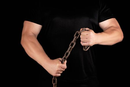 adult man in black clothes stands upright with strained muscles and holds a rusty metal chain, concept of strength and enduranceの写真素材