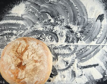round baked bread and white wheat flour scattered on a black table, top view