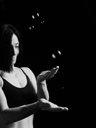 beautiful athletic woman with muscular body dressed with a black topic catches soap bubbles on a black background, studio portraits in a low keyの写真素材