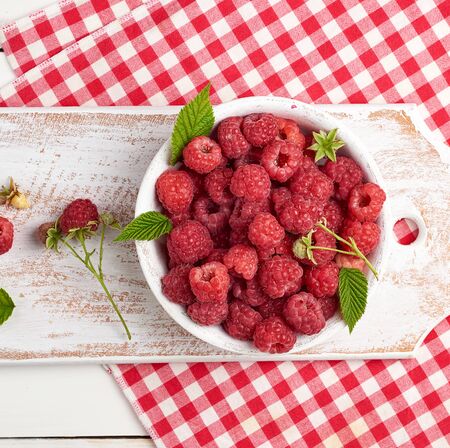ripe red raspberries in a white wooden plate on a table of boards, textile towel, top view