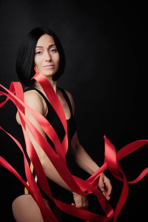 young woman gymnast of Caucasian appearance with black hair spins red satin ribbons, gymnastic exercises on black background, selective focusの写真素材