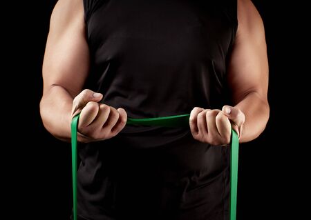 athlete with a muscular body in black clothes is doing physical exercises with green rubber, low key