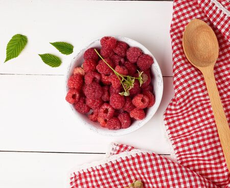 ripe red raspberries in a white wooden plate on a table of boards, next is a spoon and a textile...