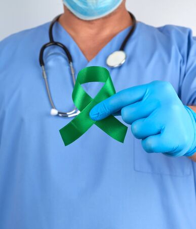 doctor in blue uniform and latex gloves holds a green ribbon as a symbol of early research and disease control,  symbol of Lyme disease, kidney transplantation and organ donationの写真素材