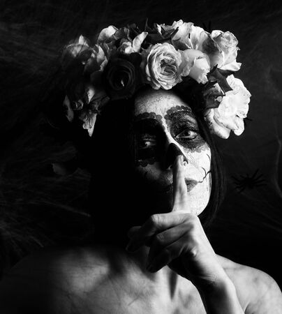beautiful girl with traditional mexican death mask. Calavera Catrina. Sugar skull makeup. girl dressed in a wreath of roses, index finger attached to lips, black and white tintingの写真素材