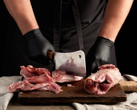 chef in black latex gloves holds a big knife and cuts into pieces raw rabbit meat on a brown wooden...