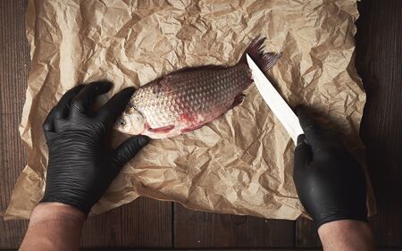 cook in black gloves cleans whole fish carp from scales on a brown piece of paper, top view