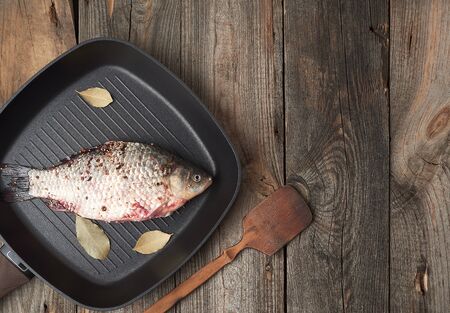 fresh crucian fish sprinkled with spices and lies in a black square pan, gray wooden table from boards, top view, copy spaceの写真素材