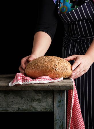 woman in a blue striped apron holds in her hands baked round rye bread and with sunflower seeds, wooden tableの写真素材