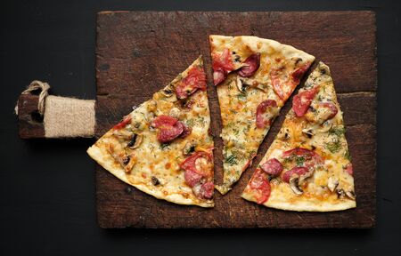 baked pizza with smoked sausages, mushrooms, tomatoes, cheese and dill, sliced food on a brown wooden board, close upの写真素材
