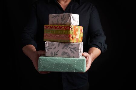 adult man in a black shirt holds in his hands a stack of paper-wrapped gifts on a black background,...